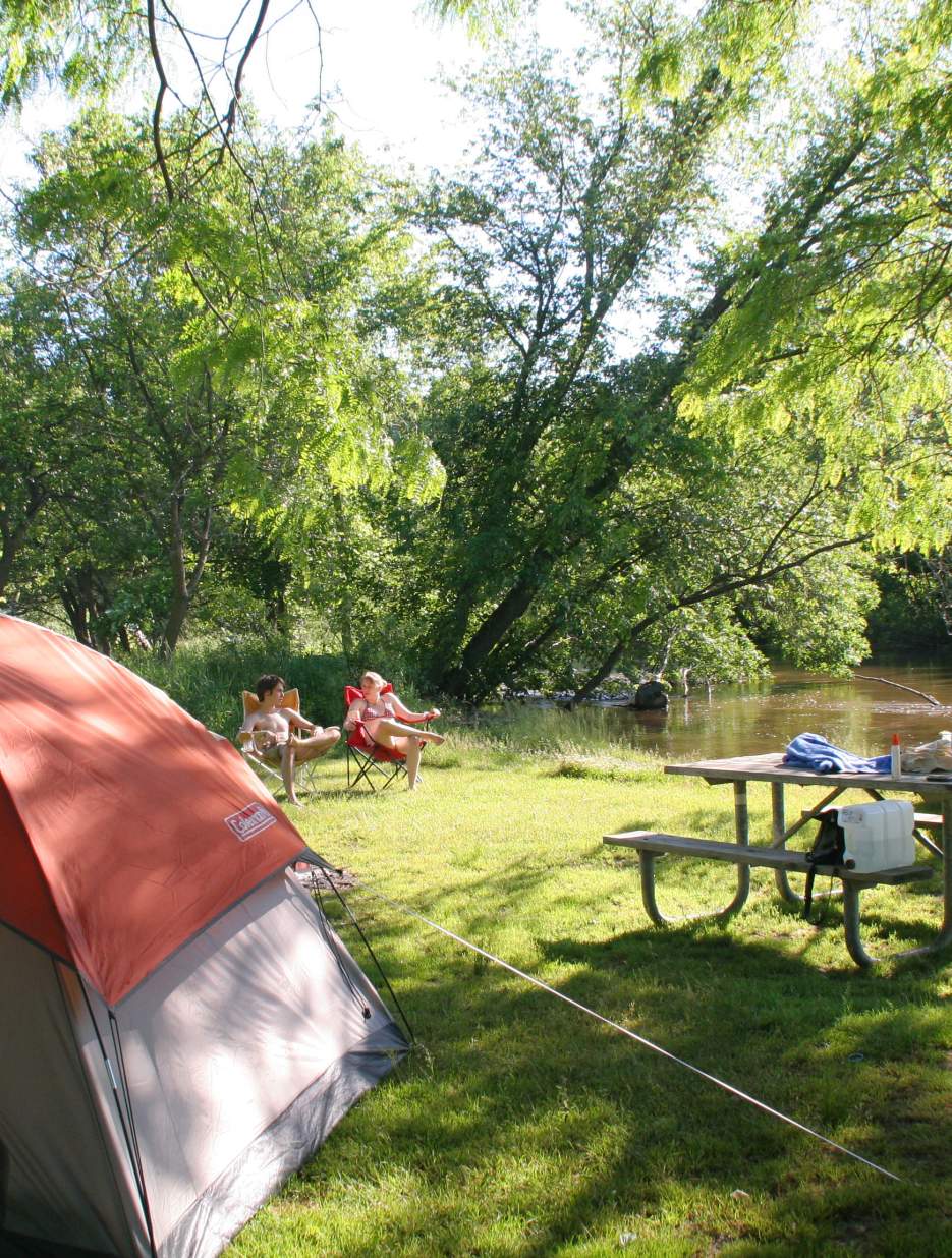 A sunny campsite by a river with a tent, picnic table, and two people relaxing in chairs, surrounded by lush green trees.