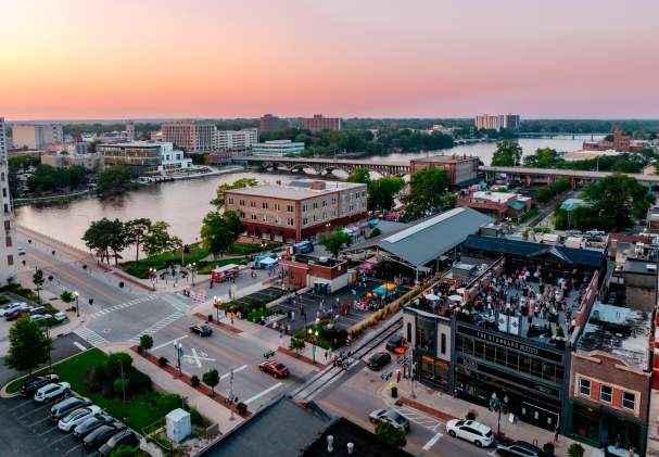 Aerial view of downtown Rockford at sunset, with people gathering near food trucks along a lively riverside, surrounded by city buildings.