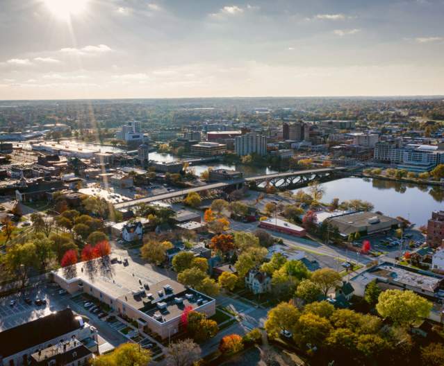 Downtown Rockford aerial view with diverse architecture, a river, and autumn foliage. Sunlight casts a warm glow over the landscape.