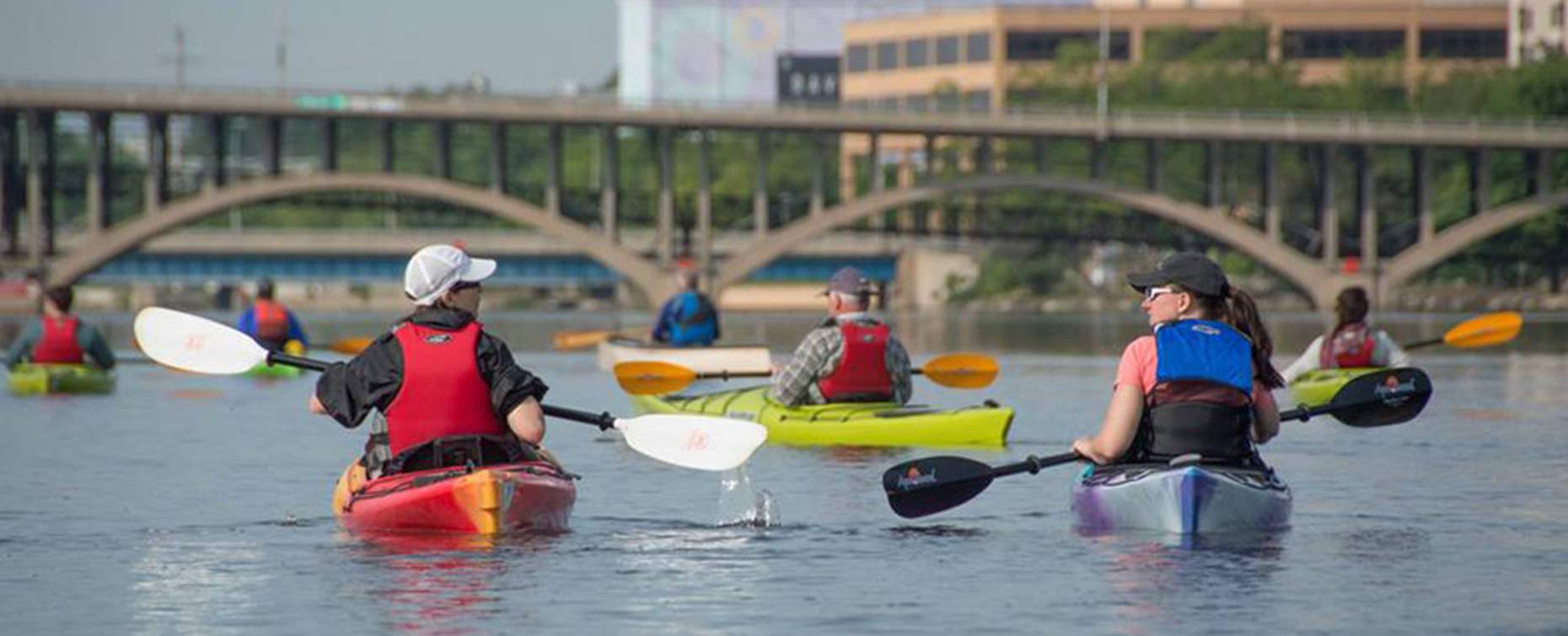 Kayakers paddling on a Rockford, IL calm river, with a bridge in the background and city buildings along the shore.