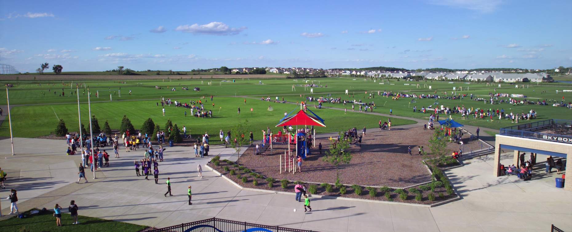 Wide view of a busy Sportscore Two complex with multiple soccer fields, playground, and people enjoying outdoor activities on a sunny day.