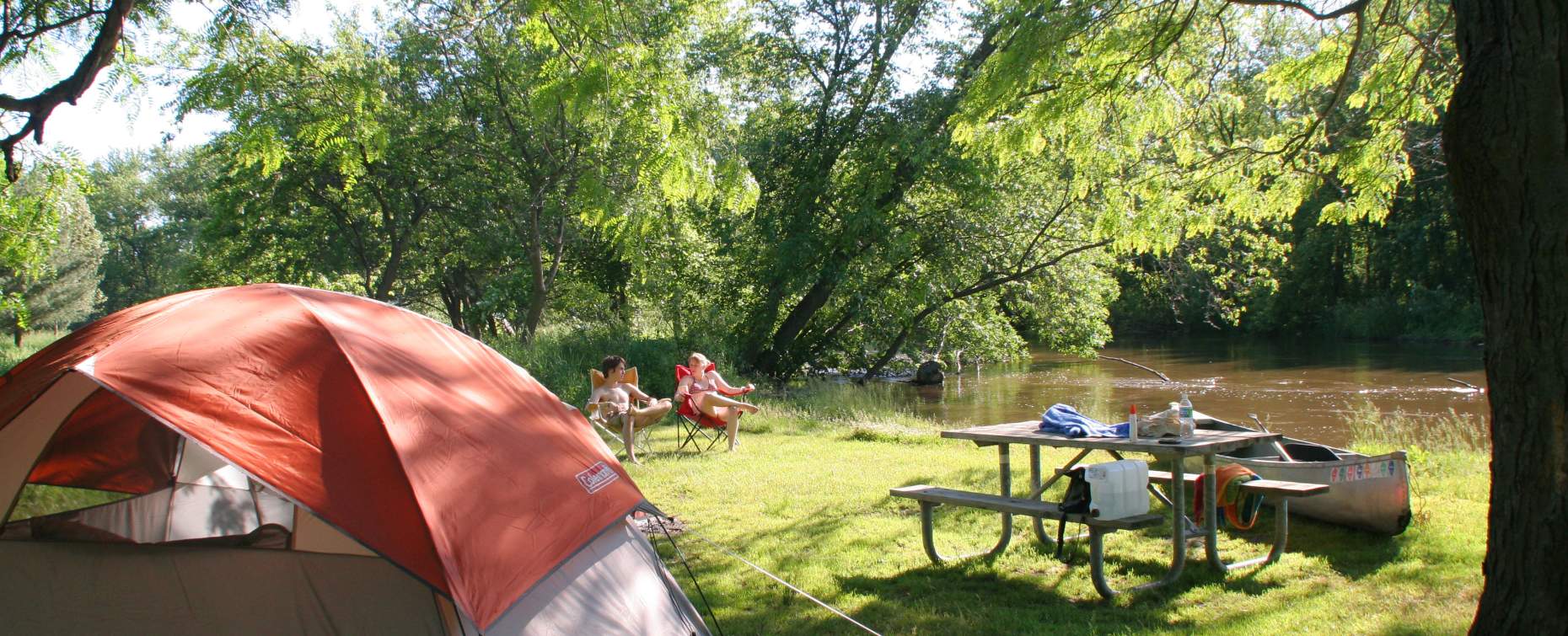 A sunny campsite by a river with a tent, picnic table, and two people relaxing in chairs, surrounded by lush green trees.