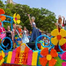Women dressed in gowns and crowns on a colorful Fiesta float in front of the Alamo.