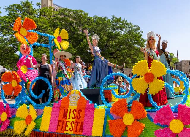 Women dressed in gowns and crowns on a colorful Fiesta float in front of the Alamo.
