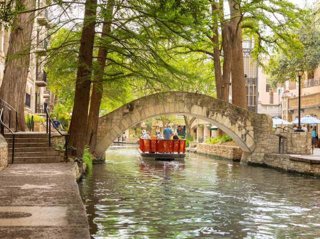 Red GO RIO River barge floating under bridge on San Antonio River Walk.