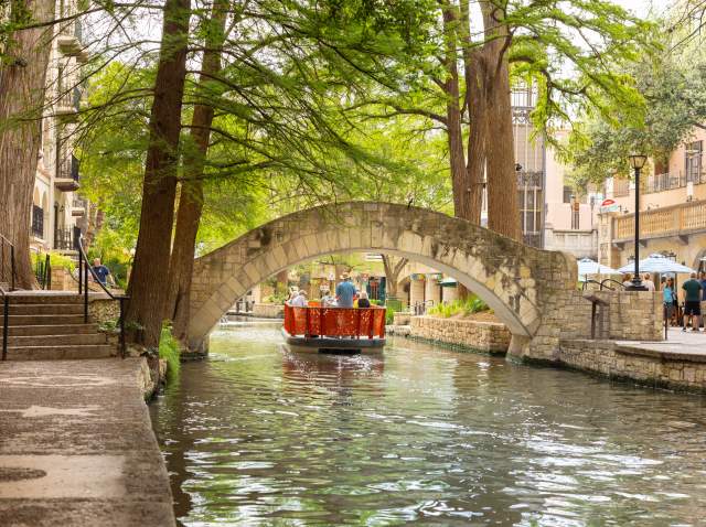 Red GO RIO River barge floating under bridge on San Antonio River Walk.