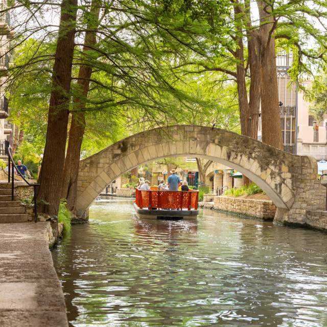 Red GO RIO River barge floating under bridge on San Antonio River Walk.
