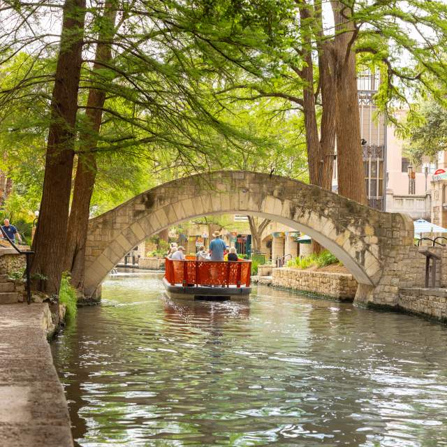 Red GO RIO River barge floating under bridge on San Antonio River Walk.