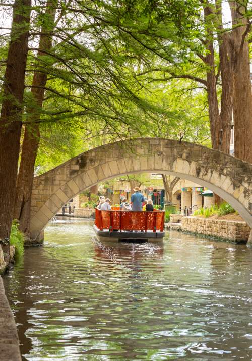 Red GO RIO River barge floating under bridge on San Antonio River Walk.