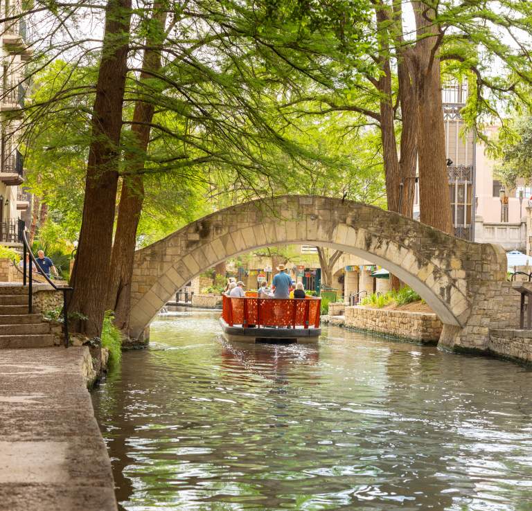 Red GO RIO River barge floating under bridge on San Antonio River Walk.
