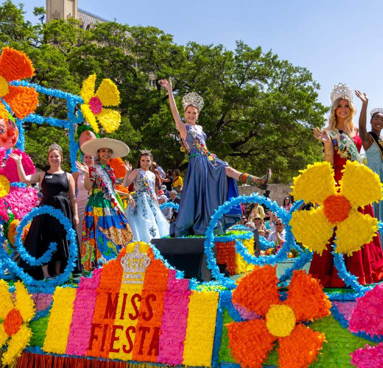 Women dressed in gowns and crowns on a colorful Fiesta float in front of the Alamo.