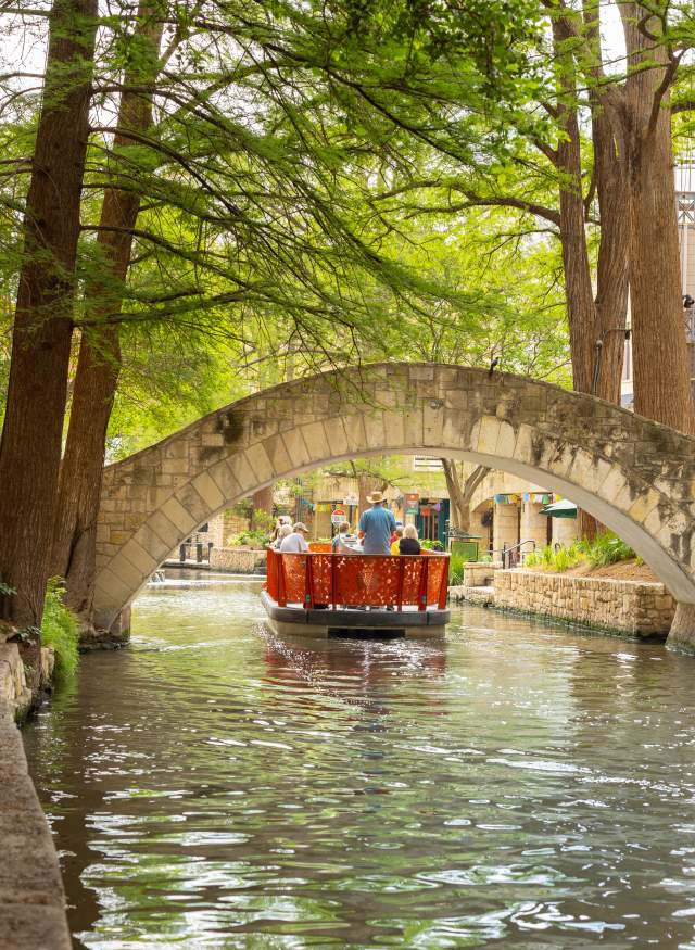 Red GO RIO River barge floating under bridge on San Antonio River Walk.