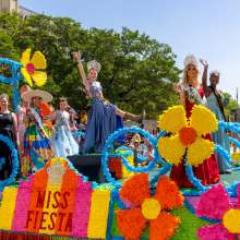 Women dressed in gowns and crowns on a colorful Fiesta float in front of the Alamo.