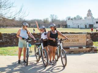 Three girls posing with bikes in front of Mission San Jose in San Antonio.