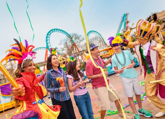 Family laughing with costumed characters at Six Flags Fiesta Texas Mardi Gras event.