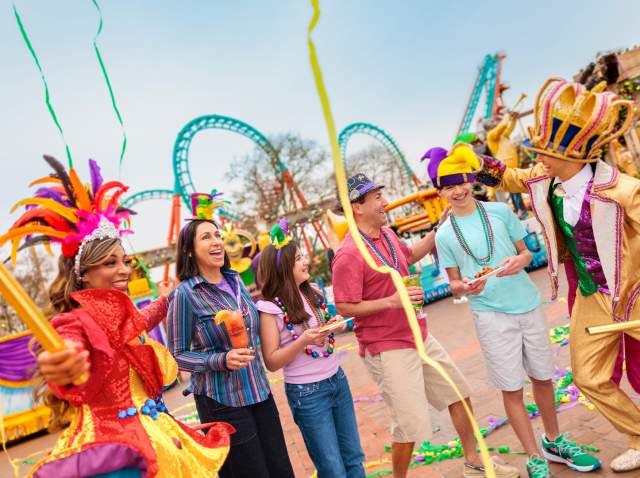 Family laughing with costumed characters at Six Flags Fiesta Texas Mardi Gras event.