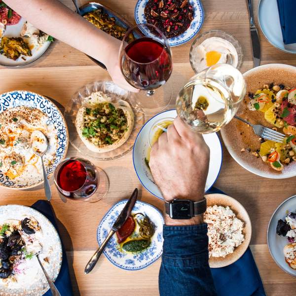 Overhead spread of food with two hands mid-toast with wine glasses