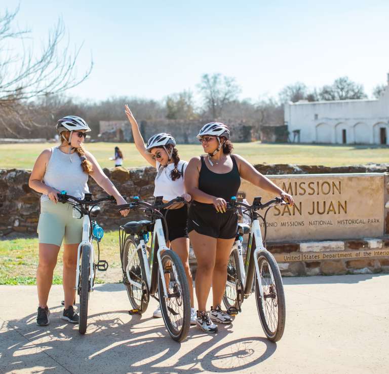 Three girls posing with bikes in front of Mission San Jose in San Antonio.
