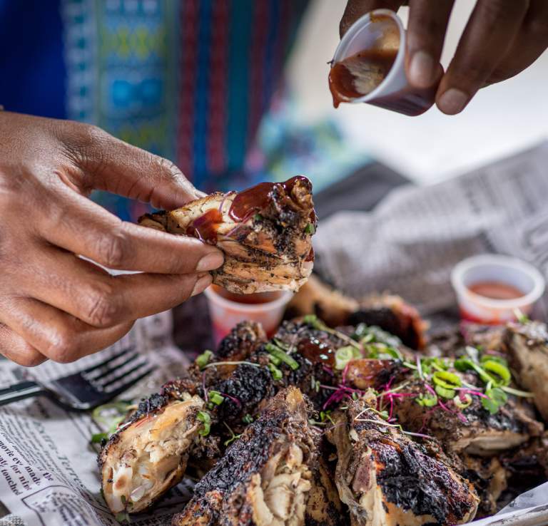 Hands dipping Jerk Chicken into barbecue sauce at The Jerk Shack.