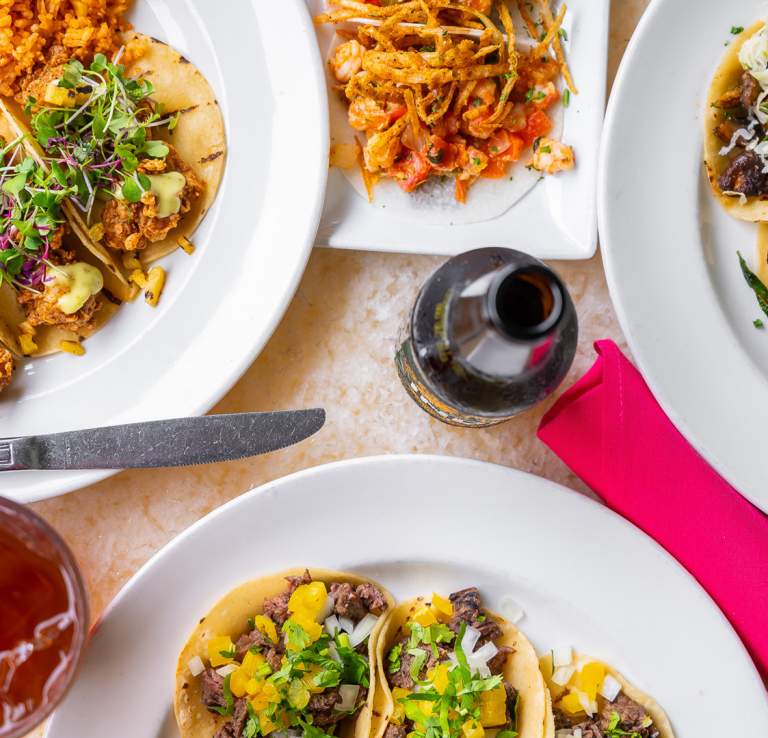 Overhead view of assorted taco plates, pink napkin, bottle, and iced tea at Acenar Restaurant.