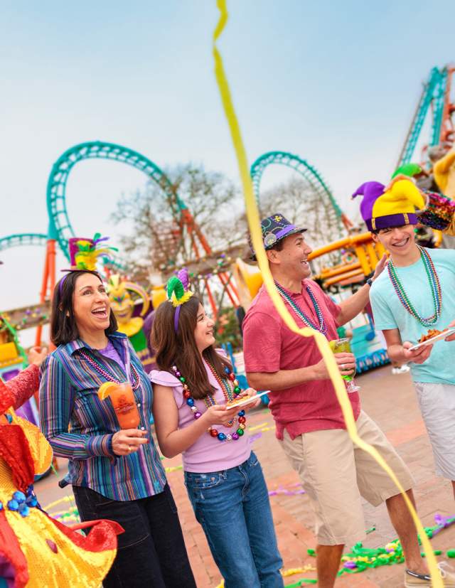 Family laughing with costumed characters at Six Flags Fiesta Texas Mardi Gras event.