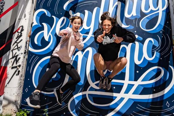 Two girls jumping in mid-air in front of street mural that reads "Everything is Magic"