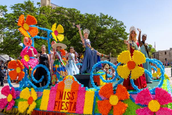 Women dressed in gowns and crowns on a colorful Fiesta float in front of the Alamo.