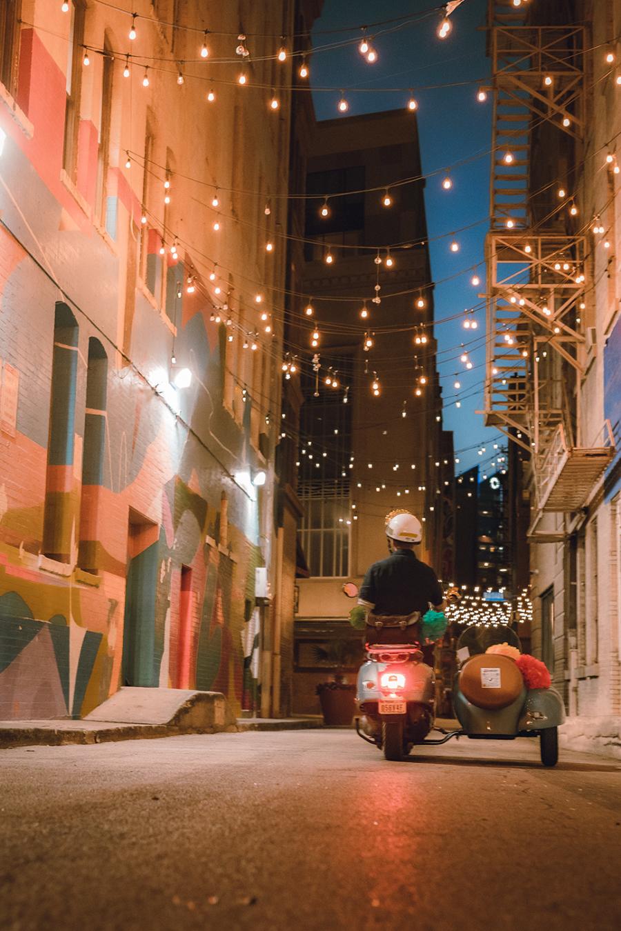 Vespa driving through alley with lights and mural on wall