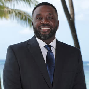 Man in suit smiling with palm tree in background.