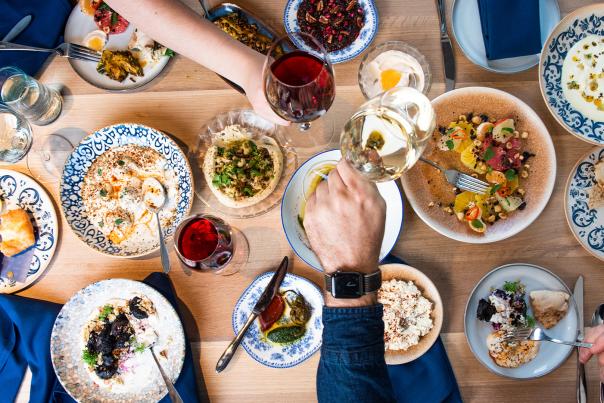 Overhead spread of food with two hands mid-toast with wine glasses