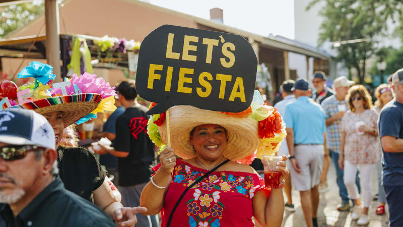 Woman wearing festive hat holding sign that says "Let's Fiesta" at Fiesta San Antonio.