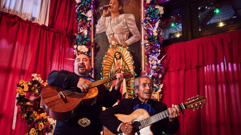 Two mariachi players in front of a painting of the Tejano artist Selena