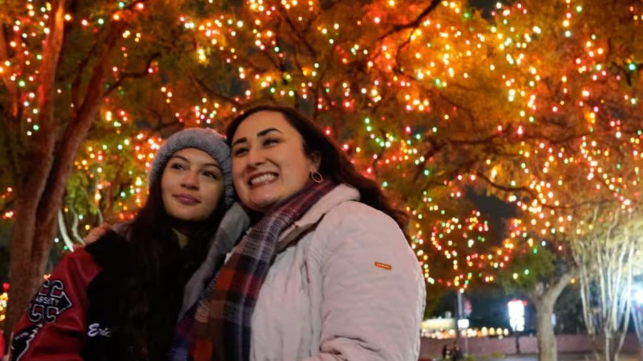 Mother and daughter posing for photo in front of tree adorned with colorful holiday lights.