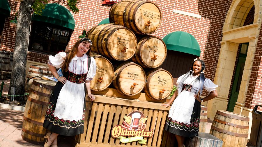 Two women dressed in traditional German attire in front of stacked barrels.