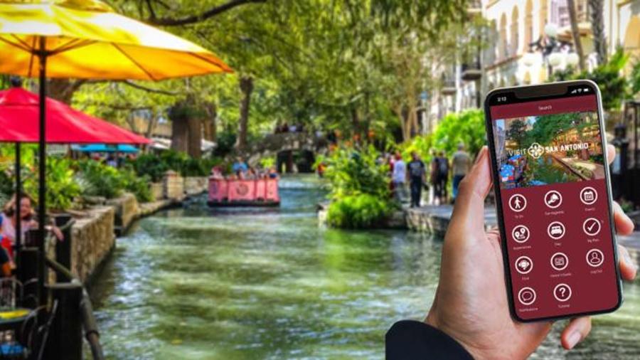 Hand holding a phone with Visit San Antonio app in front of the River Walk.