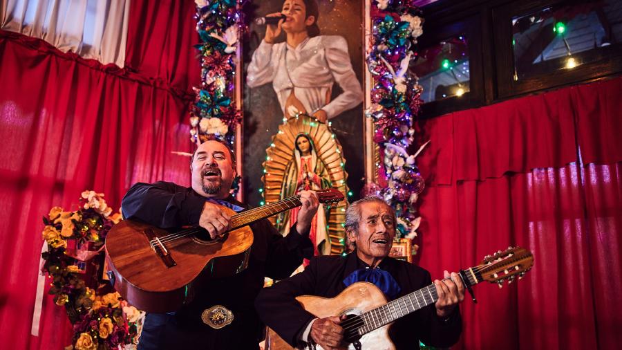 Two mariachi players in front of a painting of the Tejano artist Selena