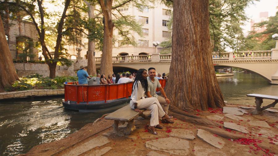 Couple sitting on bench at the River Walk in front of Go Rio RIver Cruise barge
