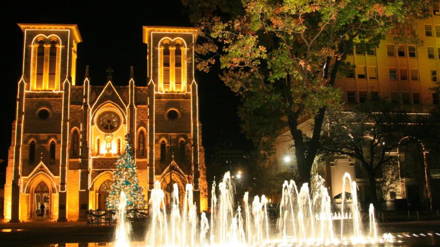 San Fernando Cathedral lit up with white Christmas lights and a lit-up water fountain in front of it.