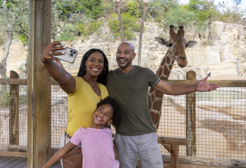 Family posing for selfie photo in front of giraffe at San Antonio Zoo.