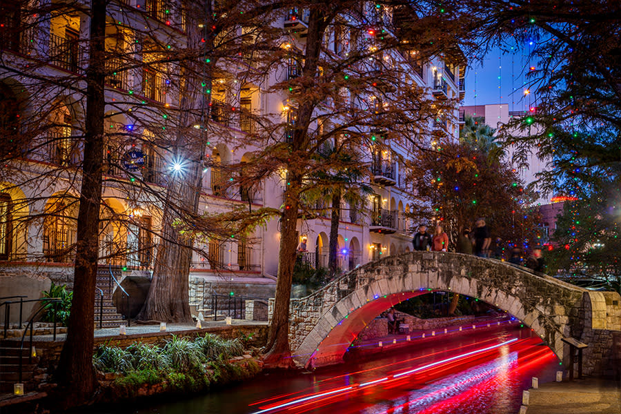 River Walk lit up with holiday lights and glow from river barge underneath bridge.