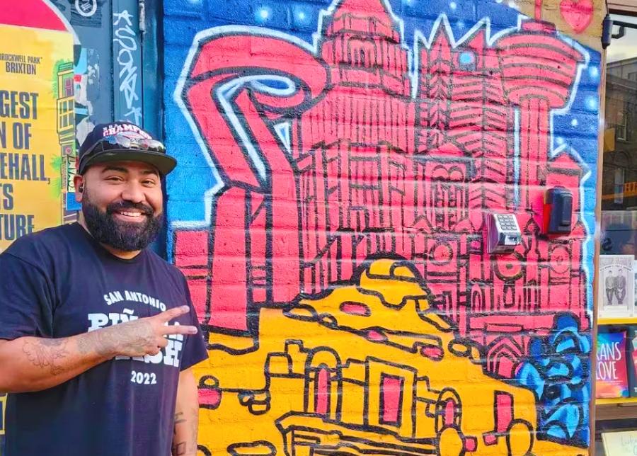 Mural artist standing in front of San Antonio mural and giving peace sign.