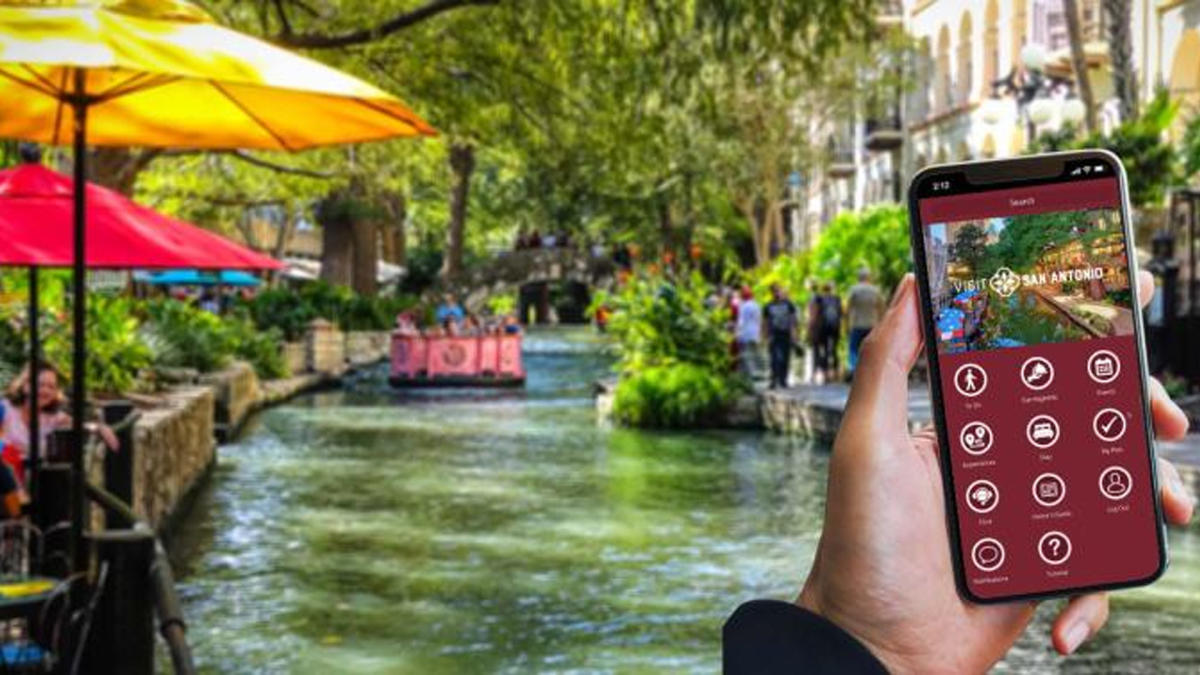 Hand holding a phone with Visit San Antonio app in front of the River Walk.