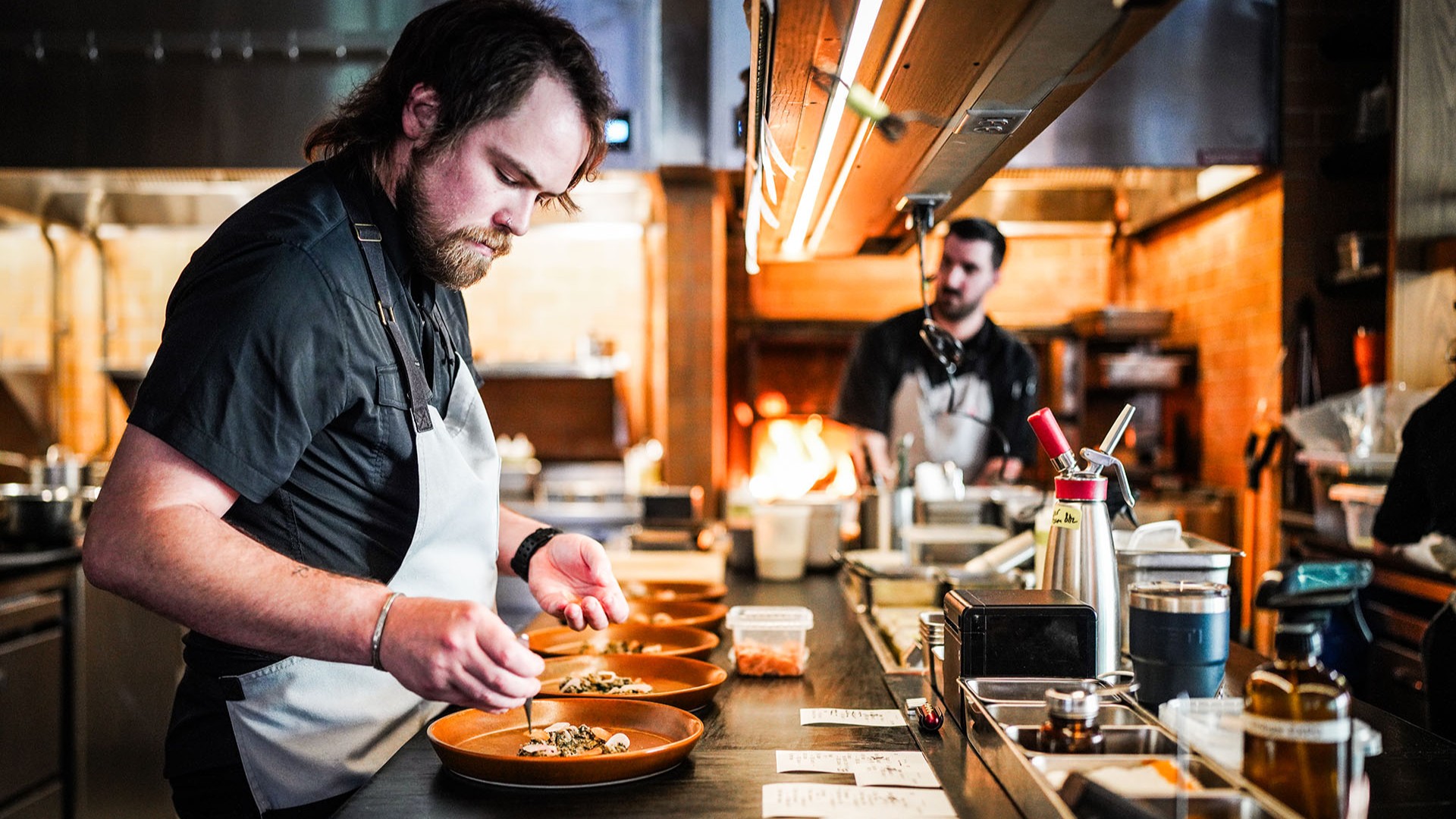 Chef putting finishing touches on dishes with another chef and fire in background.