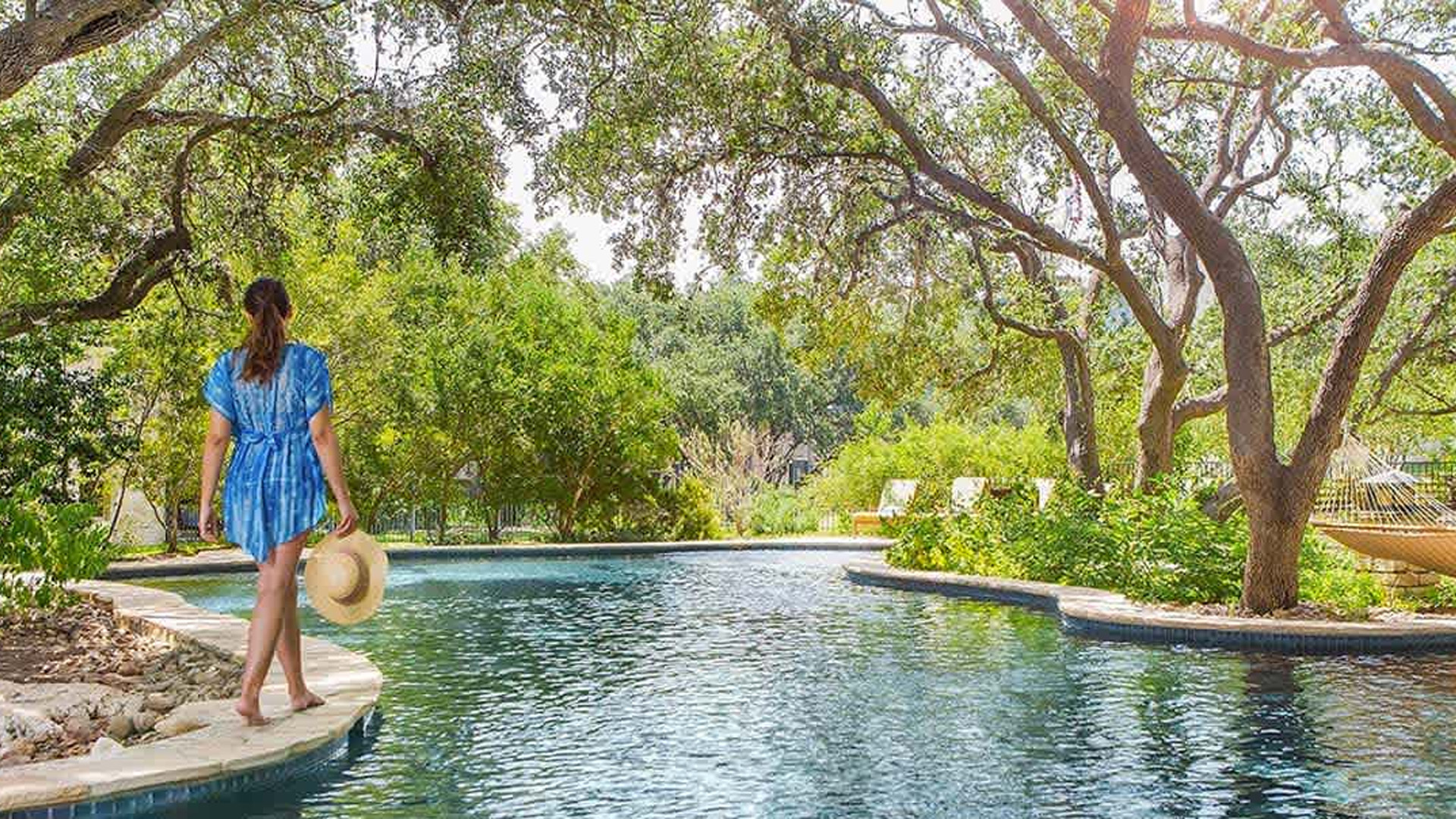 Woman walking along hotel pool holding large hat at Windflower in San Antonio.