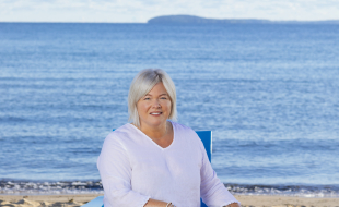 Snapshot of Traverse Tourism employee, Dana Kiel, sitting on a blue chair on the shore of Clinch Park beach in Traverse City, Michigan