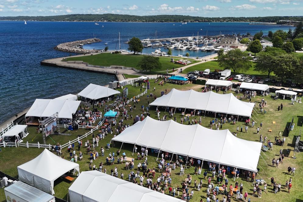An aerial view of a lawn area adjacent to the blue water of West Grand Traverse Bay with event tents set up for the Traverse City Food & Wine festival