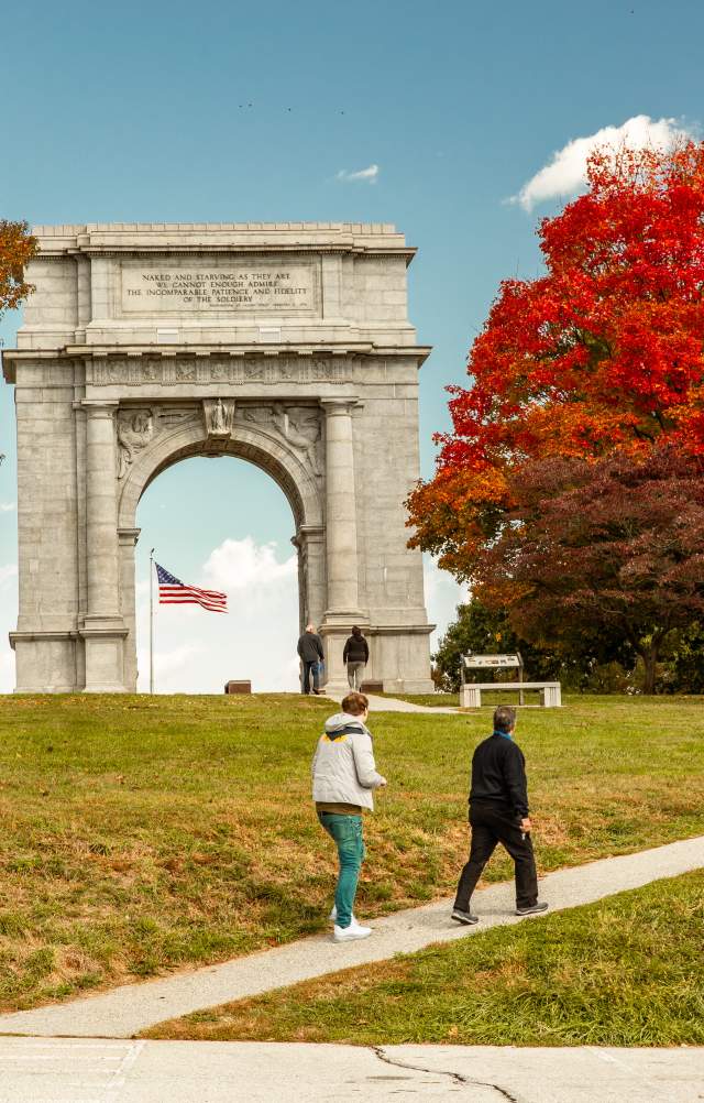 An older male and female walk casually up a hill in front of the monument arch at the Valley Forge National Historic Park surrounded by trees with fall foliage.