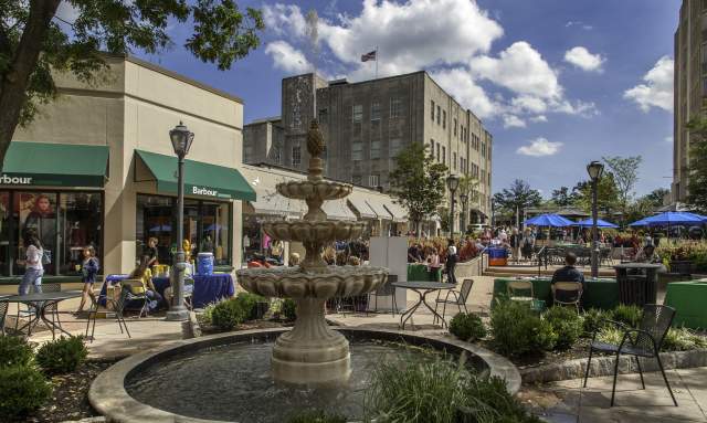 Suburban Square located in Ardmore, PA. The fountain is located in the middle of the shopping district. In the background is the storefront of Barbour. Vendors are seated at tables talking to customers. Many individuals are seen walking around enjoying the warm weather or seated outside for dining at the tables with blue umbrellas.