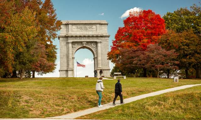 An older male and female walk casually up a hill in front of the monument arch at the Valley Forge National Historic Park surrounded by trees with fall foliage.