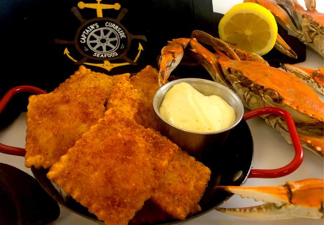 Baked ravioli with butter in a small plate with red handles is placed next to a boiled crab and sliced lemon. Behind is a shirt with the Captain's Market logo